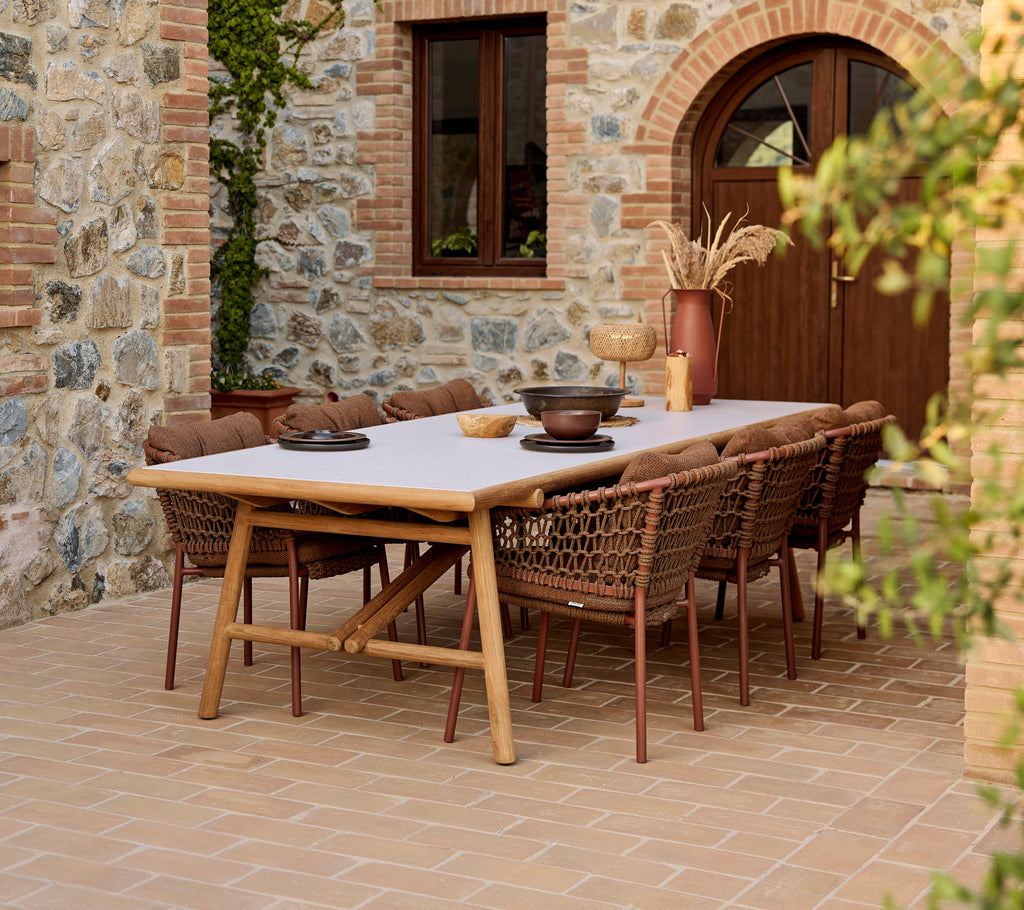 Outdoor dining setup featuring umber brown Ocean dining chairs around a teak Sticks dining table, against a stone wall backdrop.