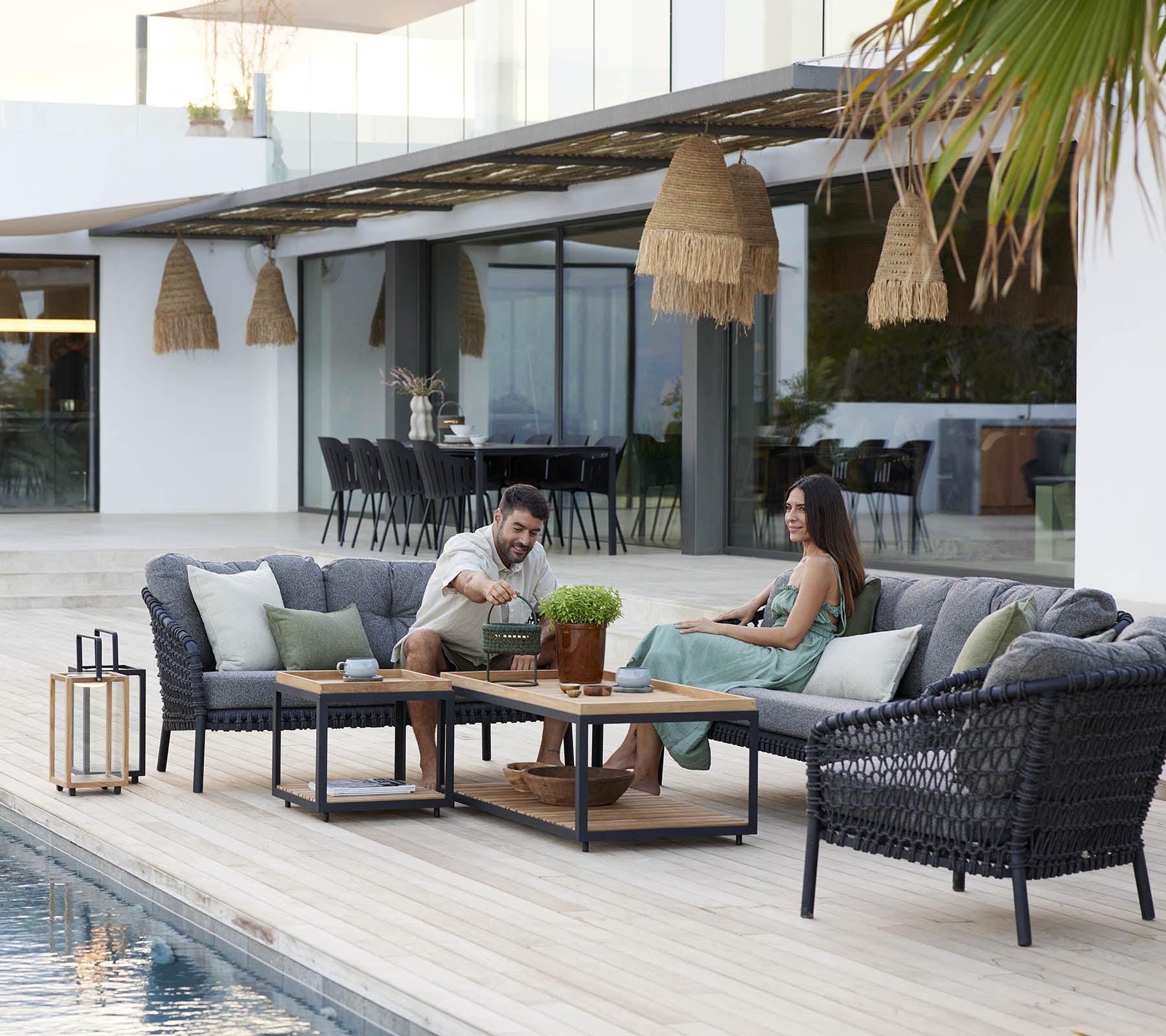 Un couple profite de son temps libre sur un ensemble de salon douillet, entouré de plantes vertes et d'une décoration élégante, avec une piscine sereine à proximité.