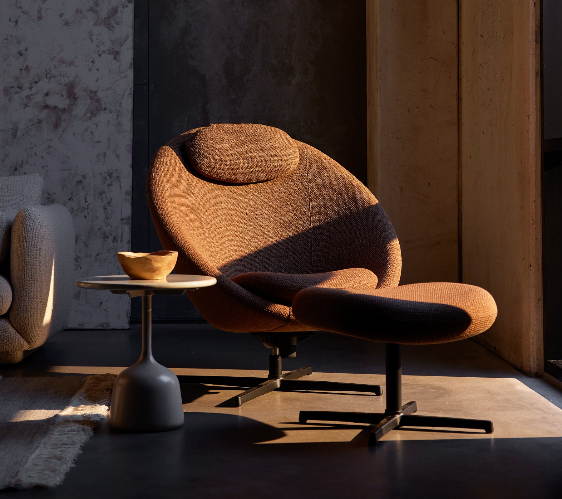 Chaise Pace brun umber avec un repose-pieds assorti, complétée par une petite table d'appoint avec un bol en bois.
