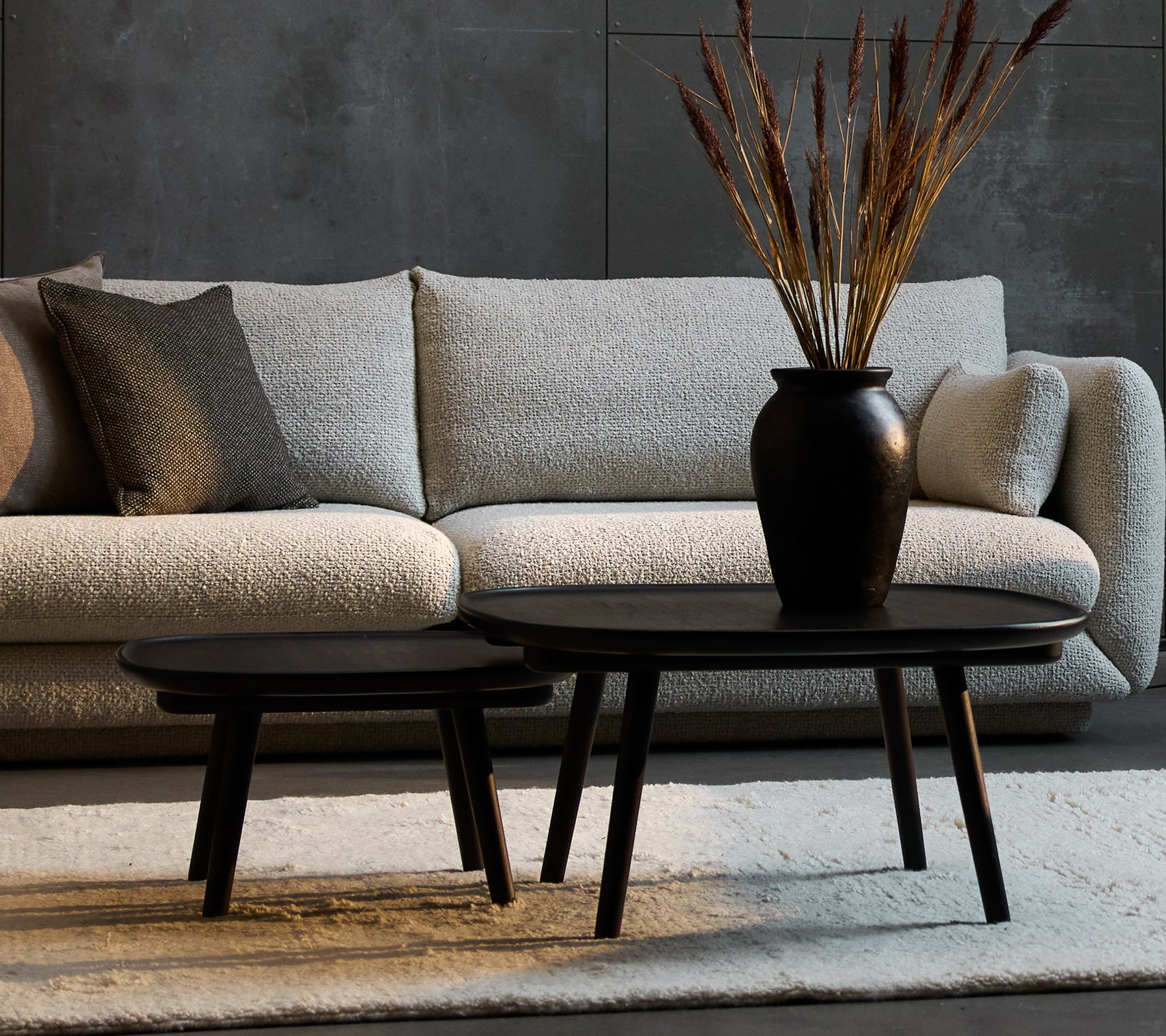 Deux élégantes tables basses noires de tailles différentes, complétées par un vase décoratif avec des plantes séchées, placées devant un canapé confortable.