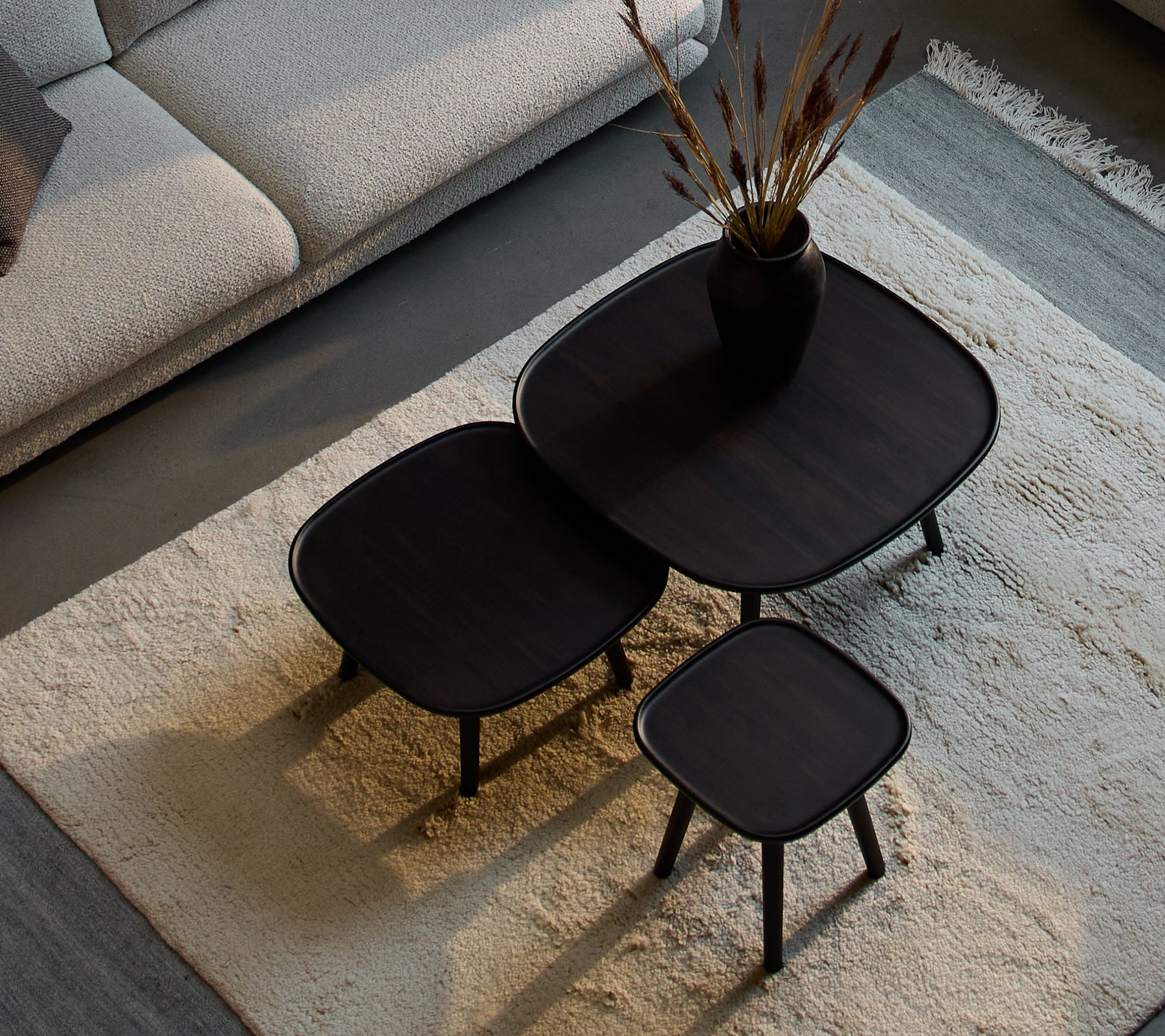 Trois tables basses Pace élégantes de différentes tailles, avec un design épuré et une finition sombre, accompagnées d'un vase décoratif et de plantes séchées.