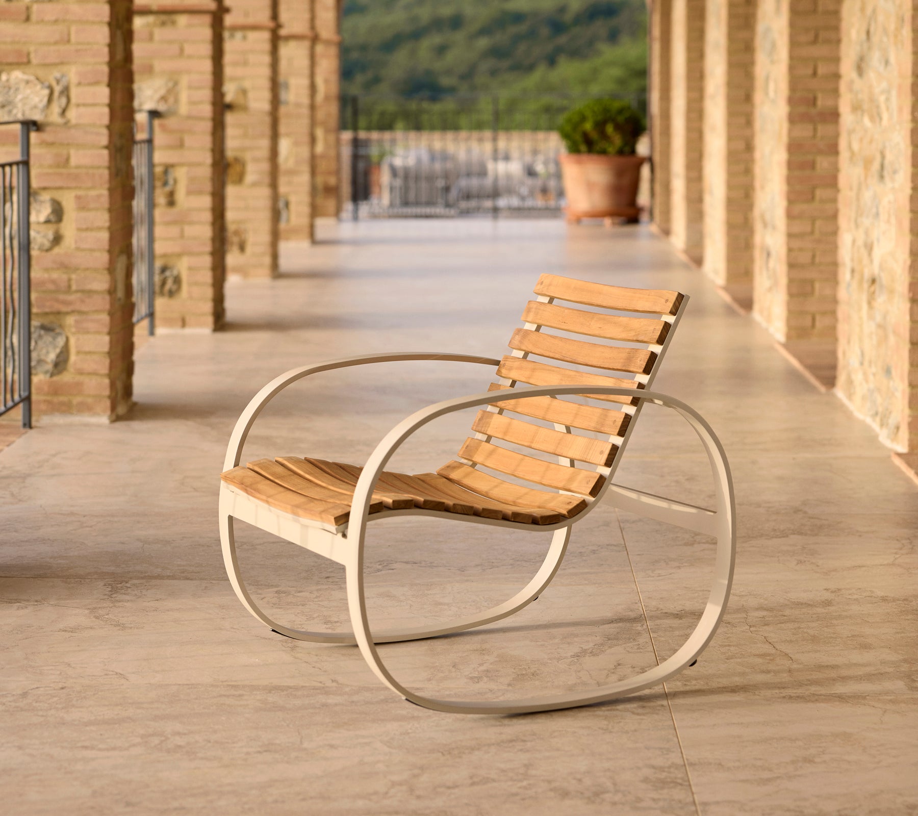 Fauteuil rocking Parc avec un cadre en aluminium et des lattes en bois, positionné sur un patio avec un sol en pierre et un cadre pittoresque.