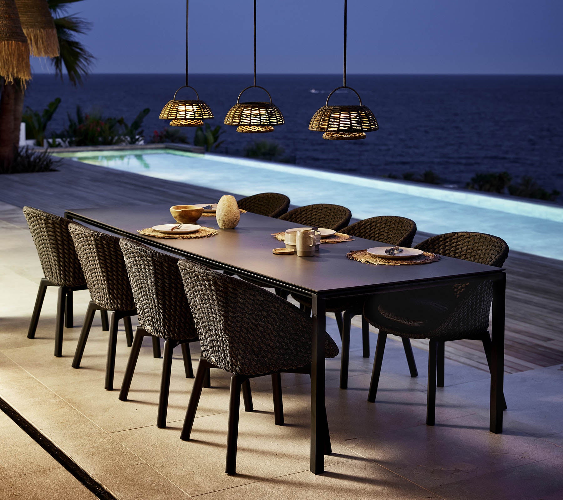Un cadre de repas élégant présente une longue table entourée de chaises paon, éclairée par des lumières suspendues au-dessus d'une vue apaisante sur l'eau.