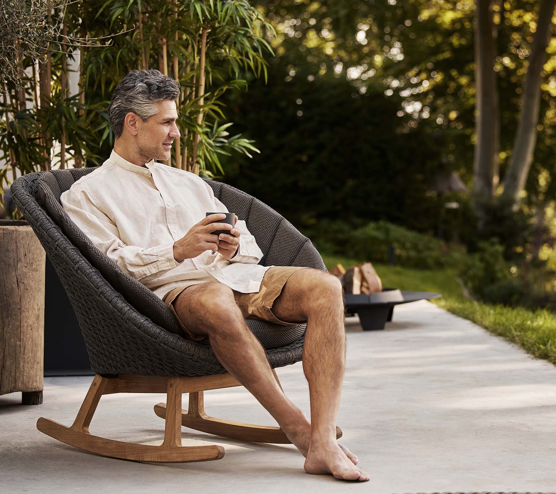 Un homme est assis confortablement dans un fauteuil à bascule en forme de paon, savourant un moment de détente tout en tenant un téléphone. De la verdure se trouve en arrière-plan.