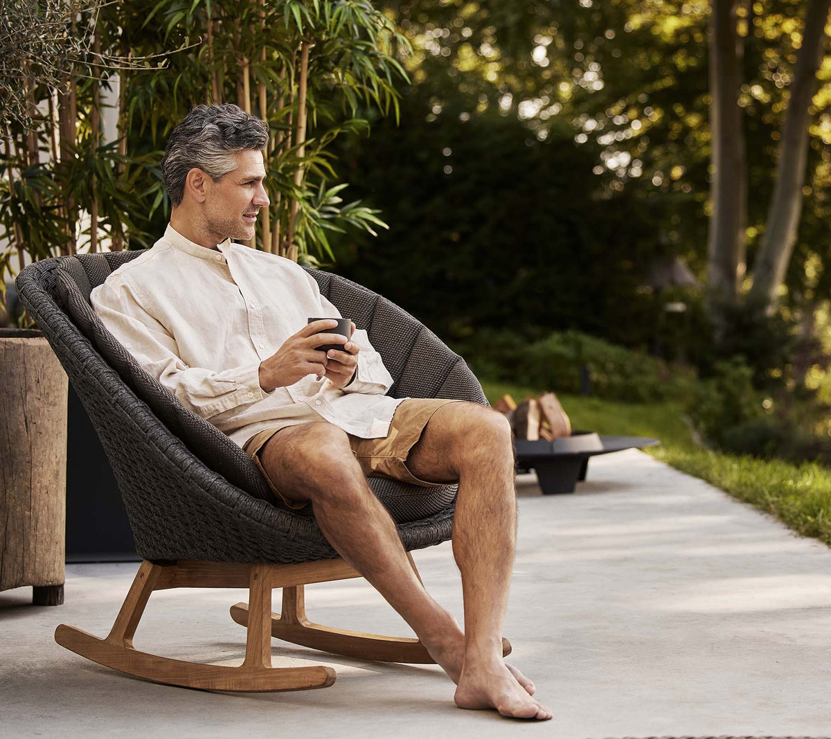 Un homme est assis confortablement dans un fauteuil à bascule en forme de paon, savourant un moment de détente tout en tenant un téléphone. De la verdure se trouve en arrière-plan.