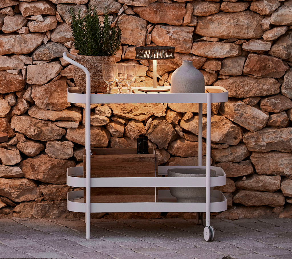 Un chariot élégant avec un dessus en bois, cadre blanc, pot décoratif et éclairage moderne contre un mur en pierre.