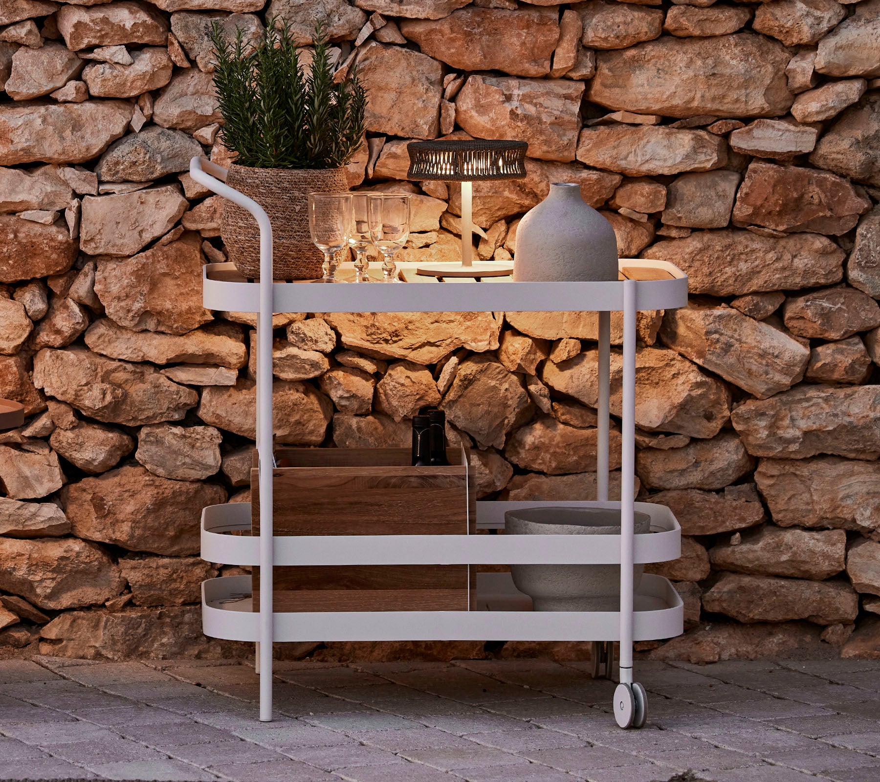 Un chariot élégant avec un dessus en bois, cadre blanc, pot décoratif et éclairage moderne contre un mur en pierre.