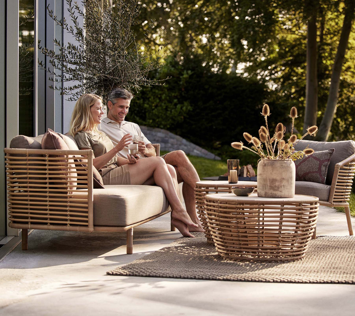 Un couple déguste des boissons sur un canapé Sense et une chaise longue, accompagné d'une table basse en osier élégante et d'un vase décoratif.