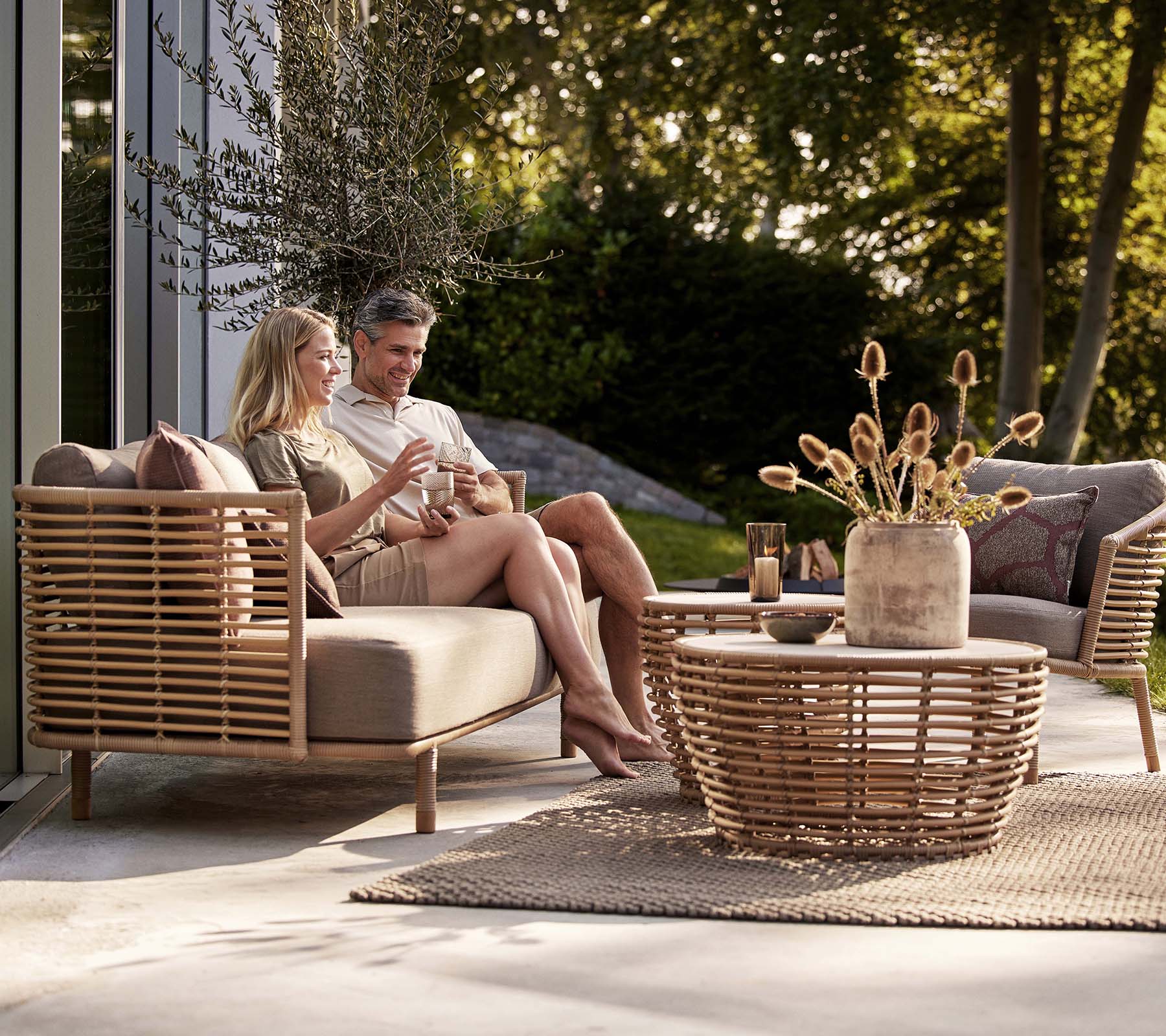 Un couple déguste des boissons sur un canapé Sense et une chaise longue, accompagné d'une table basse en osier élégante et d'un vase décoratif.