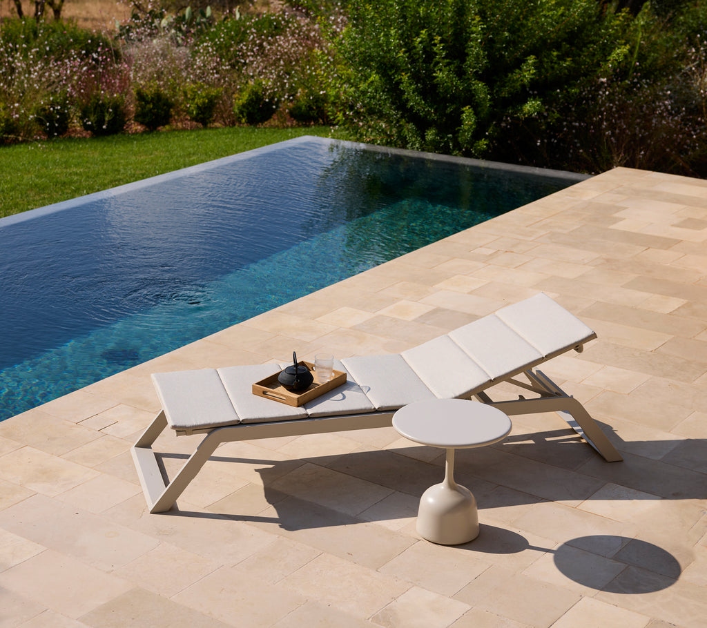 Chaise longue blanche moderne et table d'appoint au bord d'une piscine entourée de verdure.