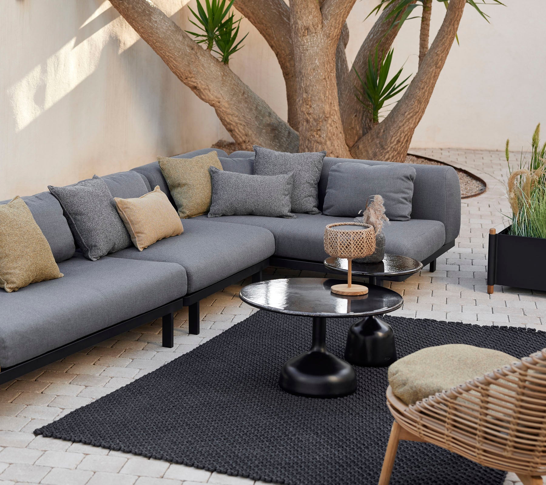 Espace salon avec un canapé modulaire gris avec des coussins décoratifs, une table basse épurée et un tapis texturé entouré de verdure.