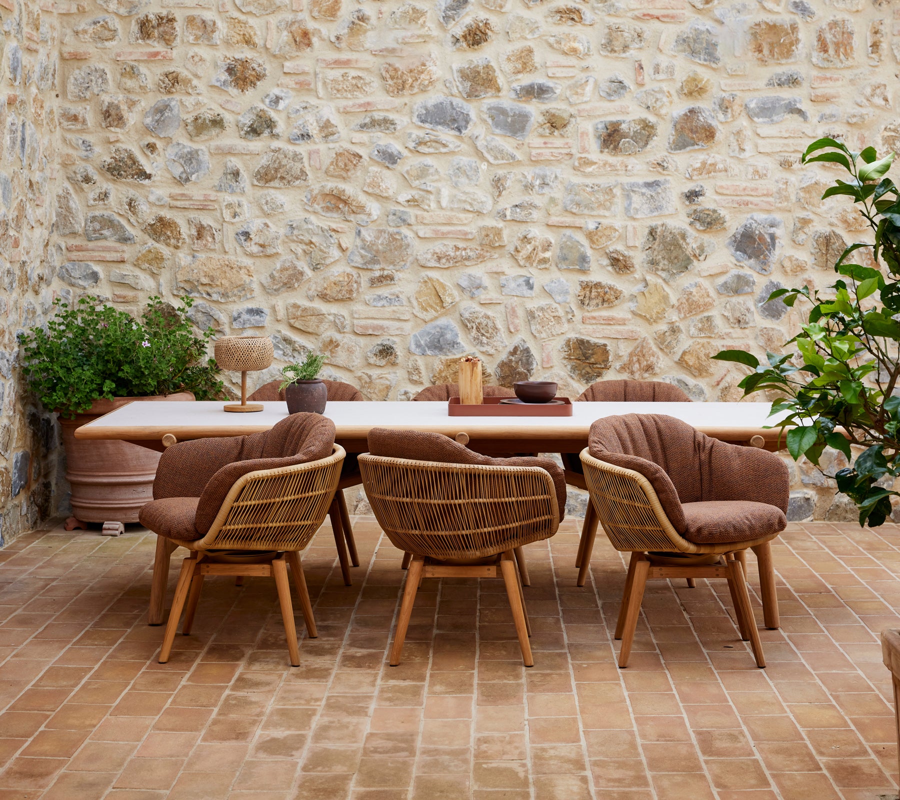 Une table à manger en bois entourée de chaises élégantes avec des pieds en teck, contre un mur en pierre texturé et ornée de plantes vertes.