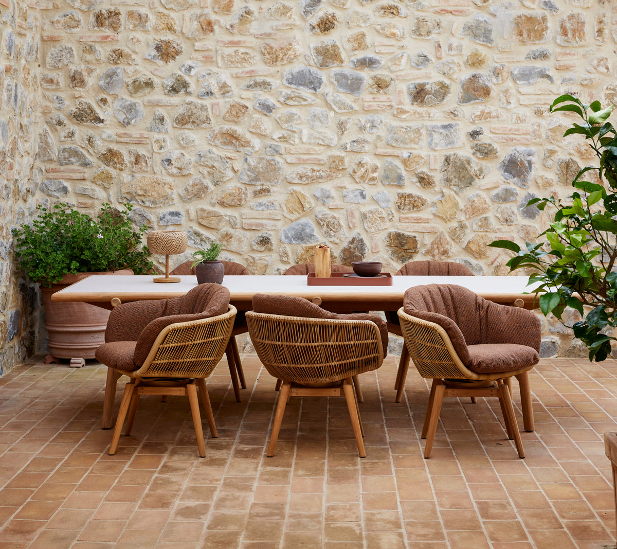 Une table à manger en bois entourée de chaises élégantes avec des pieds en teck, contre un mur en pierre texturé et ornée de plantes vertes.