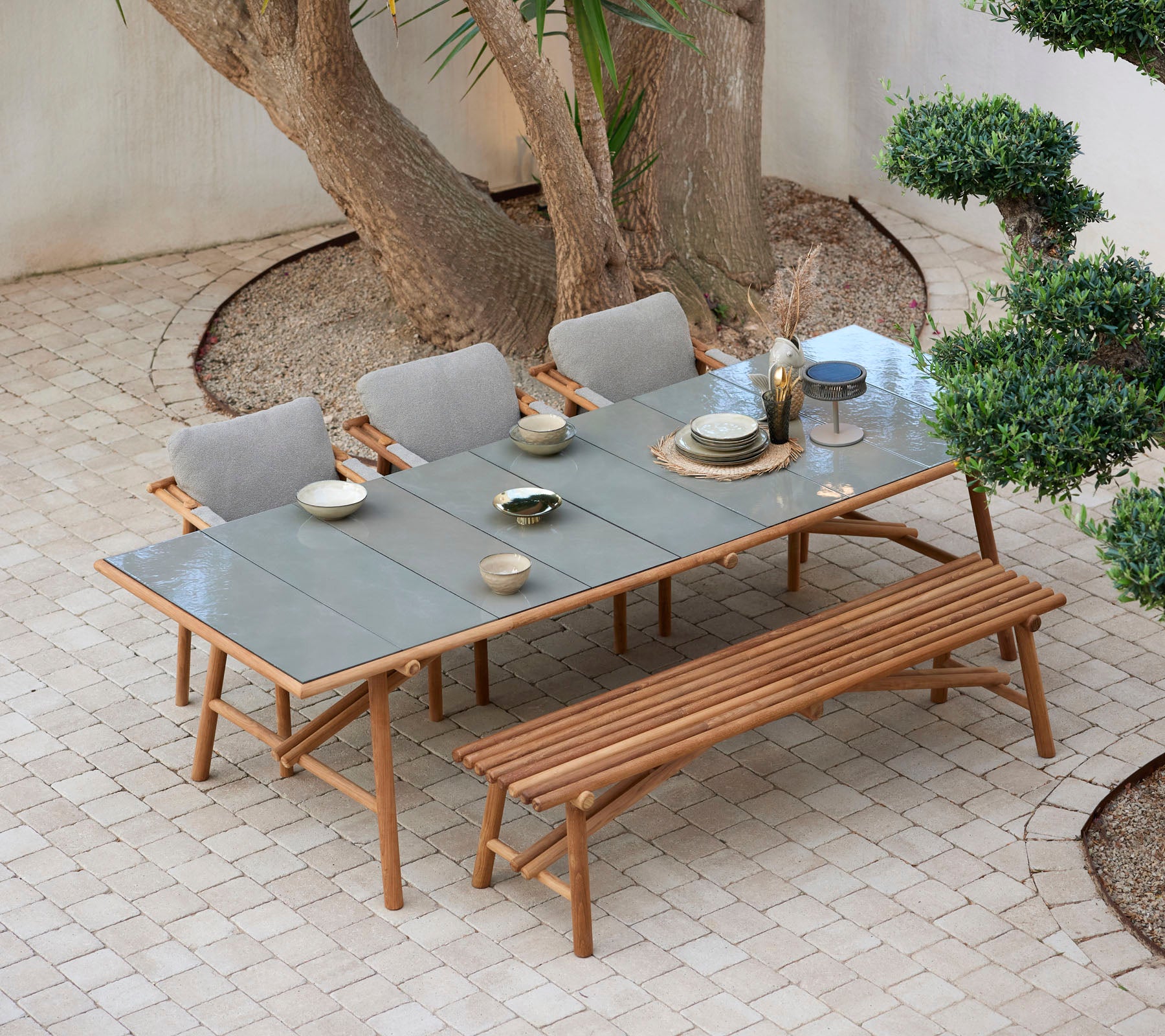 Une table en bois dressée avec des assiettes et des verres, entourée de chaises confortables et d'un banc, le tout positionné parmi la verdure.