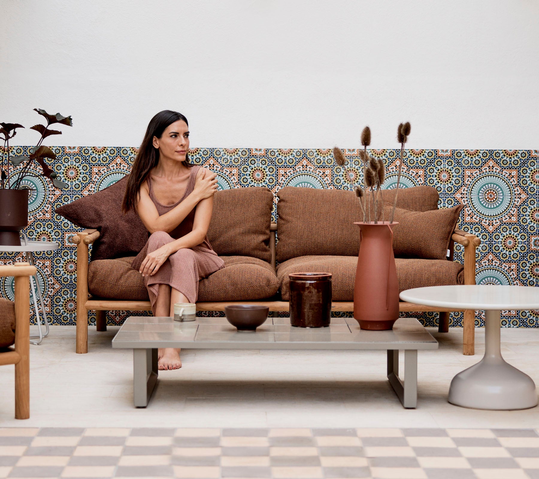 Une femme assise sur un canapé avec des coussins décoratifs, entourée de vases et de plantes, contre un fond à motifs.