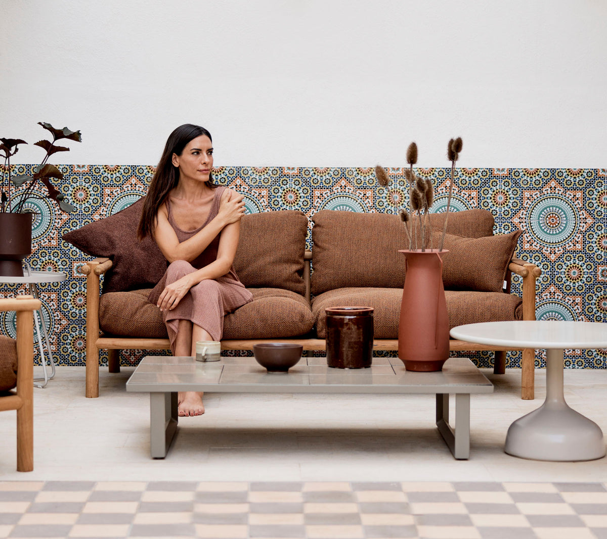 Une femme assise sur un canapé avec des coussins décoratifs, entourée de vases et de plantes, contre un fond à motifs.