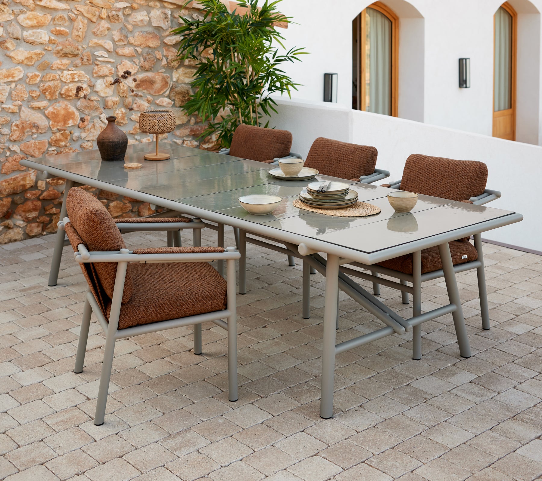 Un ensemble de table à manger spacieux avec des bâtons en aluminium, des chaises rembourrées brunes et de la vaisselle disposée sur une surface en verre.