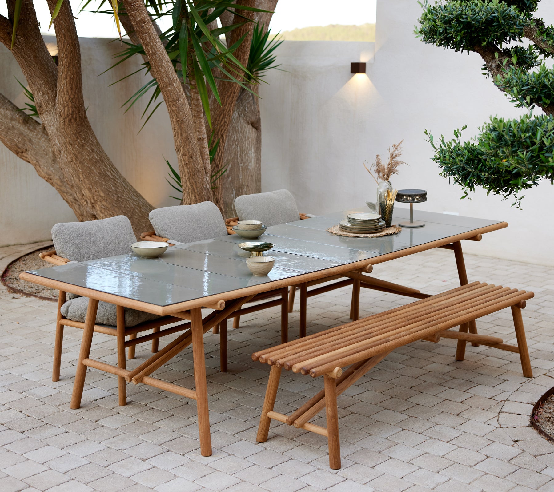 Table à manger en teck avec dessus en verre, accompagnée d'un banc et de quatre chaises rembourrées, dressée avec des plats et un éclairage d'ambiance.