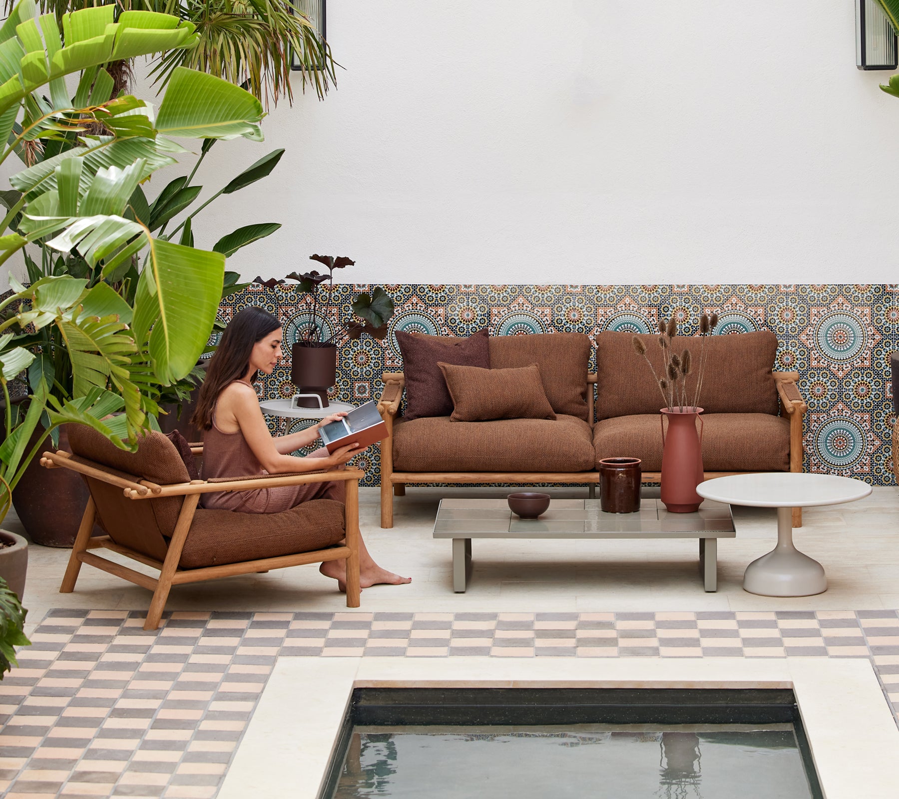 Une personne lisant un livre dans un coin salon douillet avec des meubles en teck, des coussins, une table basse et de la verdure environnante.
