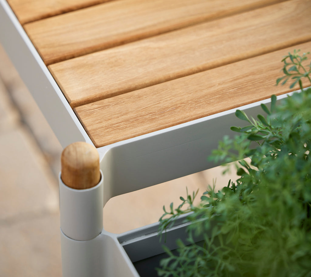 Banc en teck avec un plateau à lames, un cadre blanc et des plantes vertes, comportant un détail en bâton en bois arrondi.