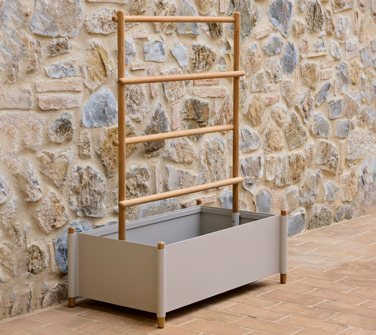 Un pot en aluminium gris avec un support en bois, conçu pour cultiver des plantes, placé contre un mur en pierre texturé.