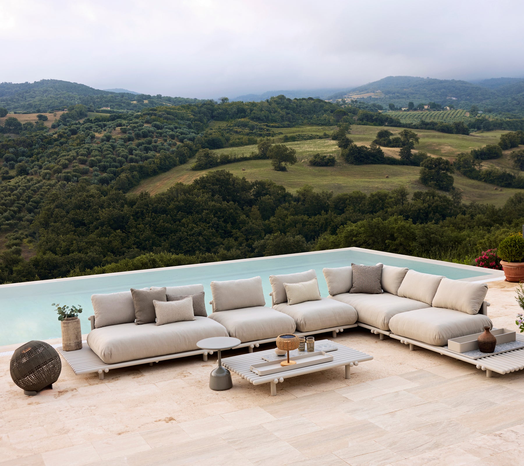 Mobilier lounge en aluminium sur une plateforme, avec des coussins neutres et une lampe Illusion ronde, dans un paysage pittoresque.