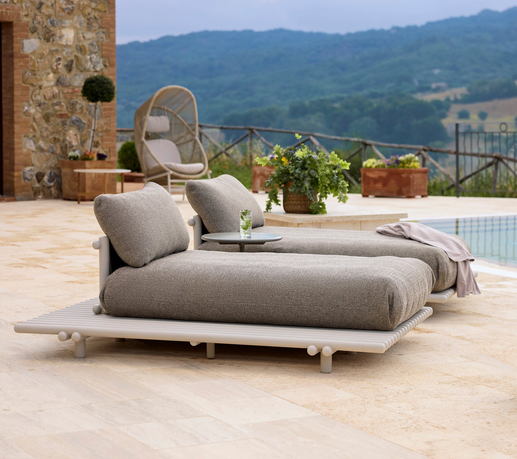 Chaises longues en aluminium élégantes avec de douces coussins, accompagnées d'une petite table, sur fond de verdure et de collines pittoresques.
