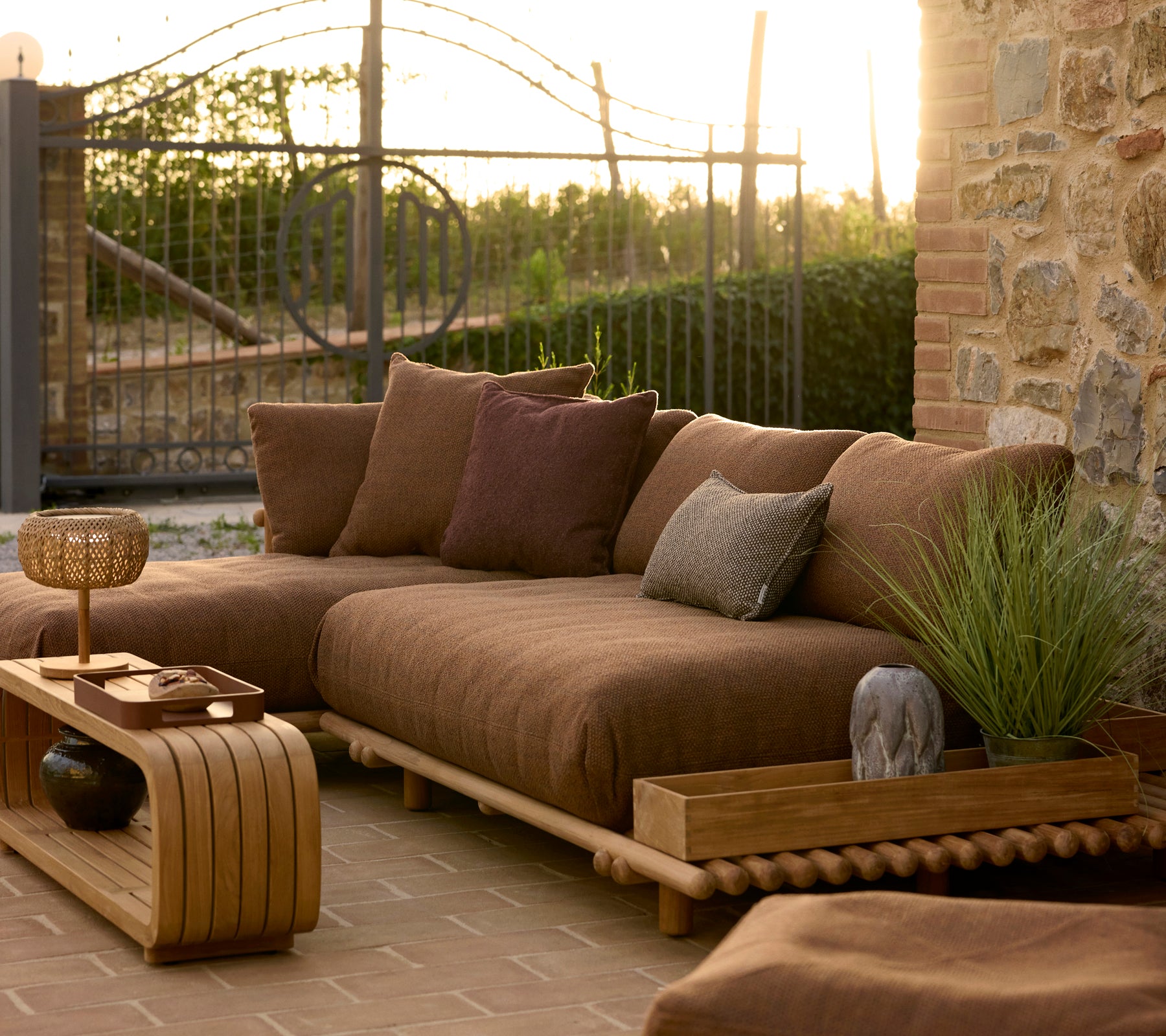 Table basse décontractée et boîte de rangement en teck à côté d'un coin salon extérieur confortable avec des coussins et des plantes sous la lumière douce du soir.