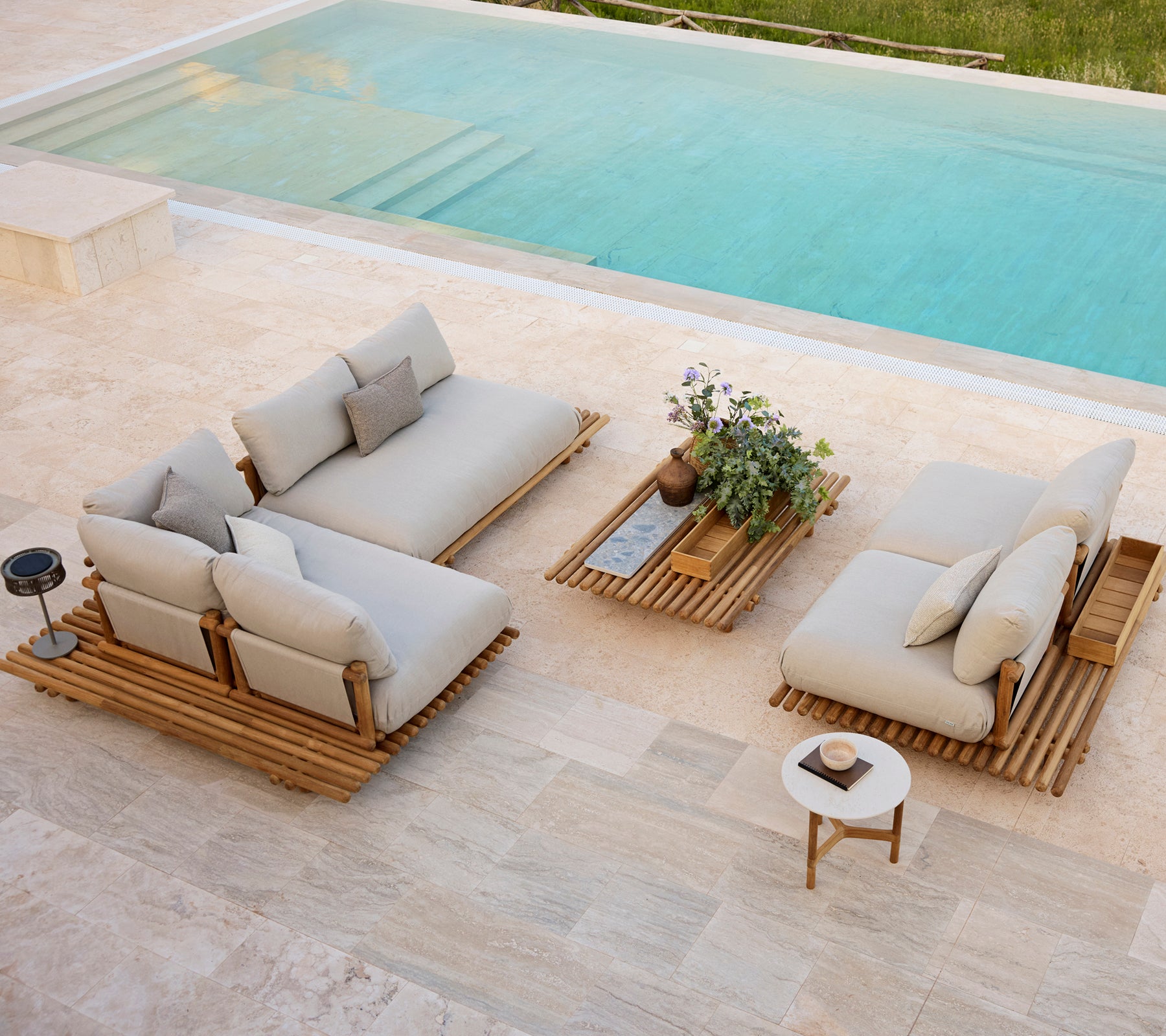 Espace extérieur moderne avec des coussins neutres, des tables en bois et une petite plante en pot à côté d'une piscine.