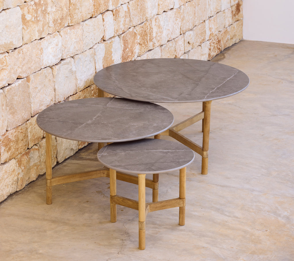 Trois tables basses grises modernes avec des bases en bois clair contre un mur en pierre.