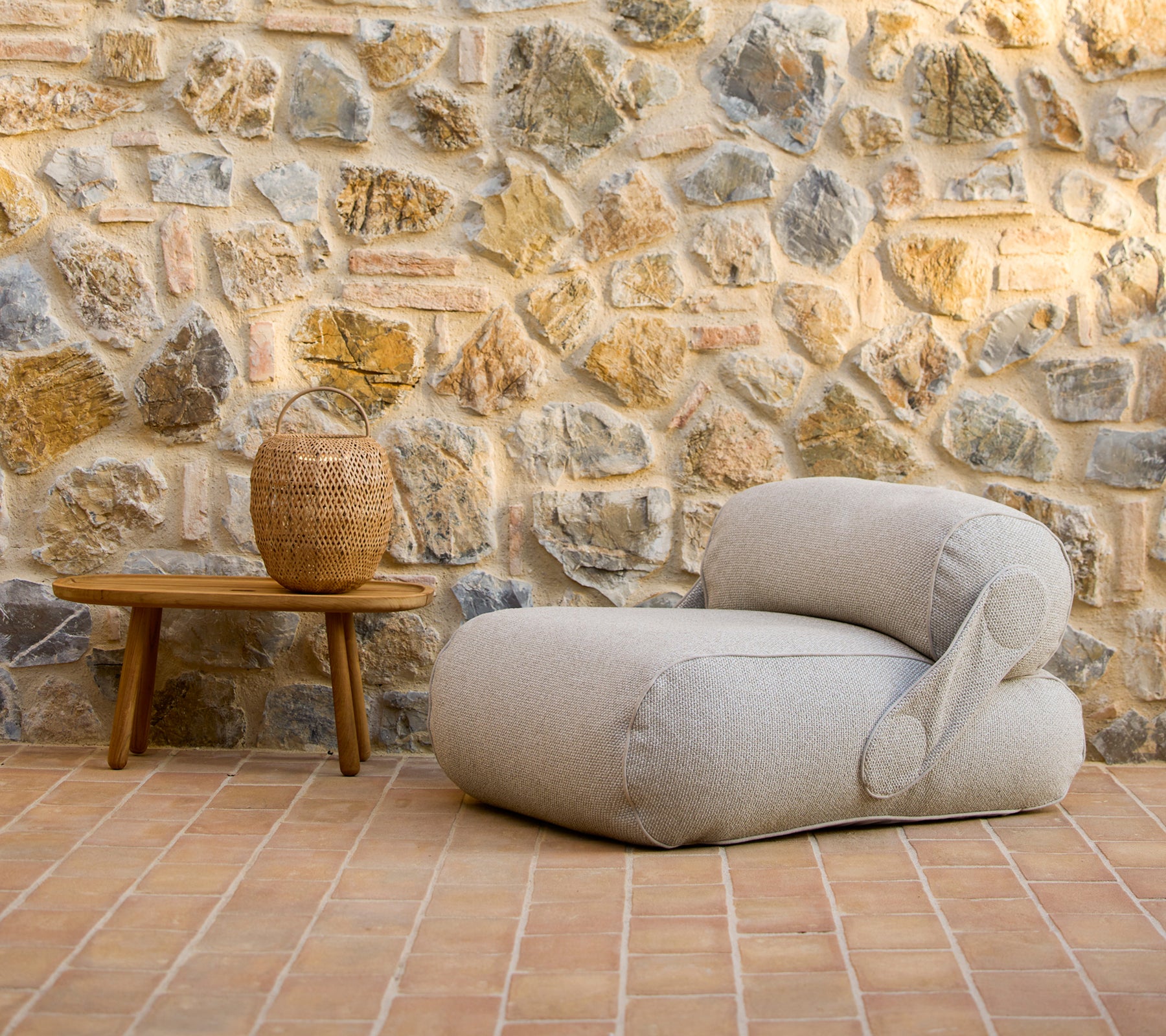 Un fauteuil lounge élégant à côté d'une table basse en bois et d'un vase décoratif, contre un mur en pierre rustique.