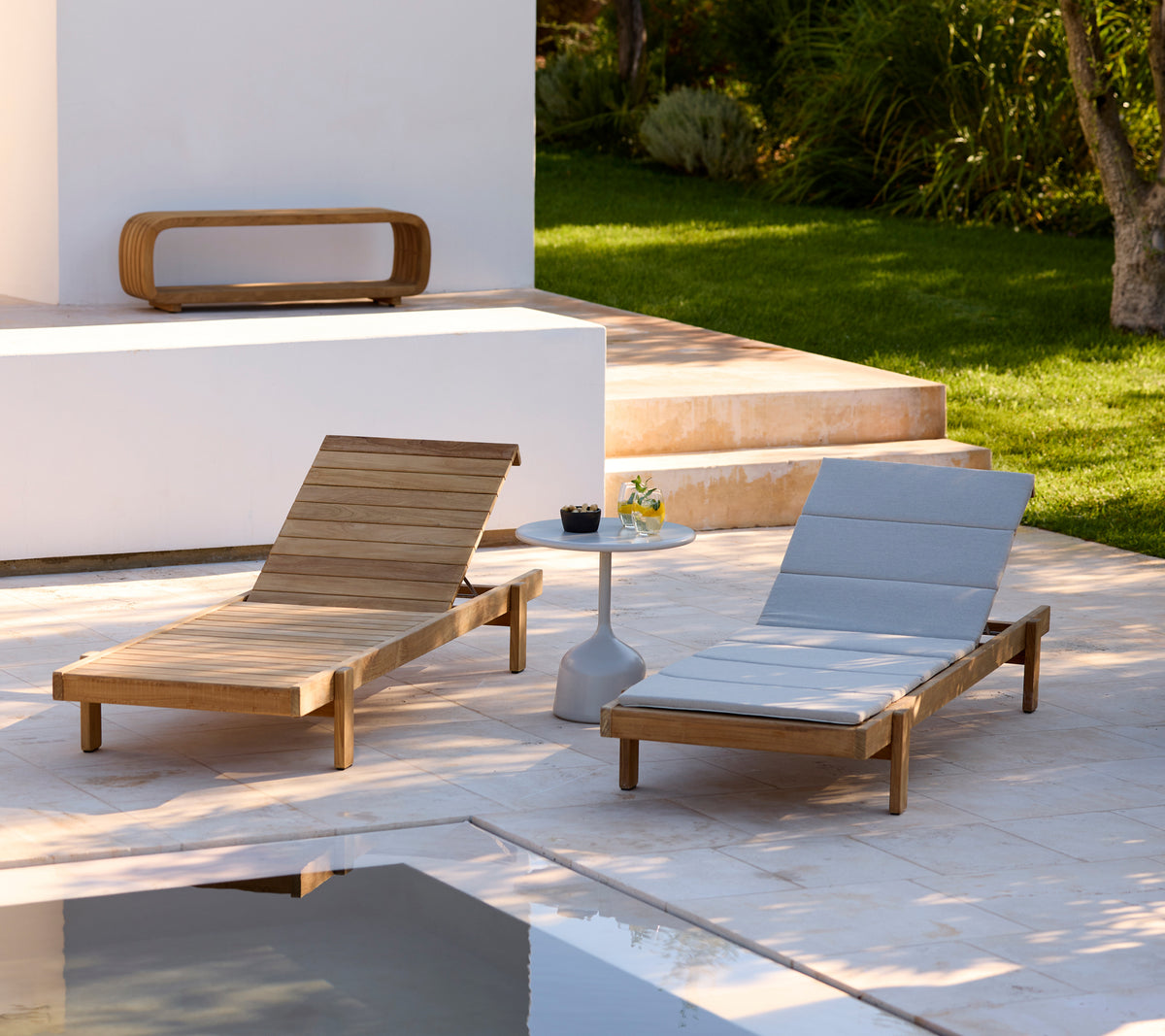 Deux chaises longues en bois modernes avec des coussins gris et une table d'appoint blanche dans un cadre extérieur.