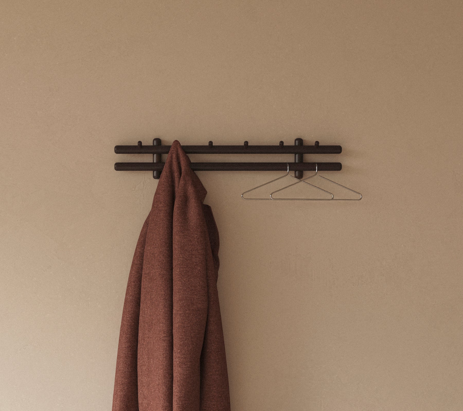 Un manteau marron pend au-dessus d'un porte-manteau mural minimaliste avec des crochets, complété par un simple cintre en fil contre un fond neutre.