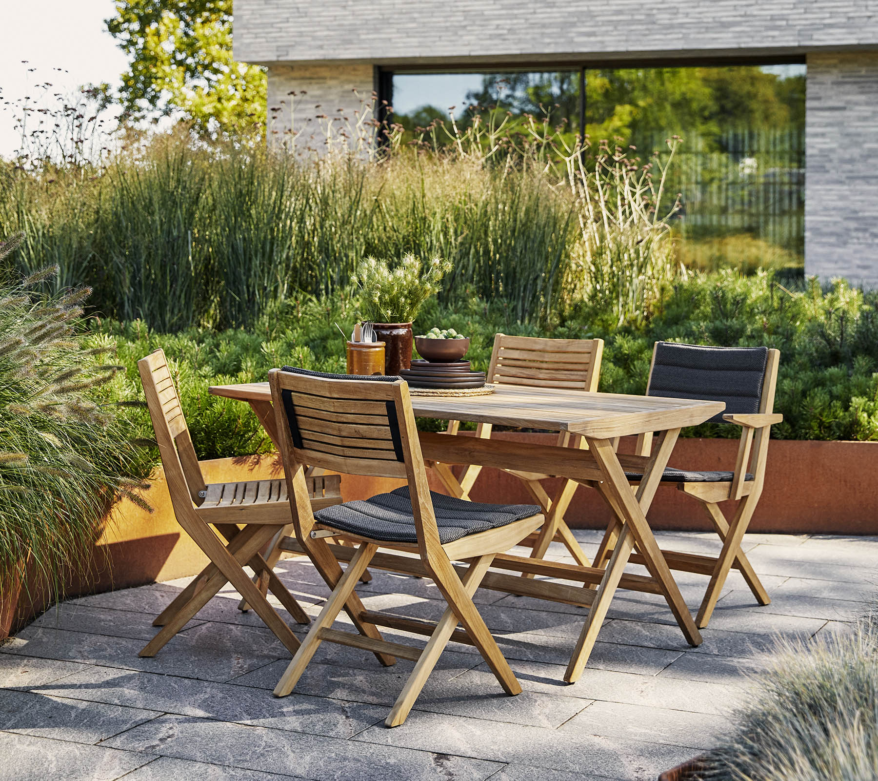 Table en bois élégante avec des chaises assorties entourées d'une verdure luxuriante et d'un arrière-plan architectural moderne. Parfait pour des rassemblements en extérieur.