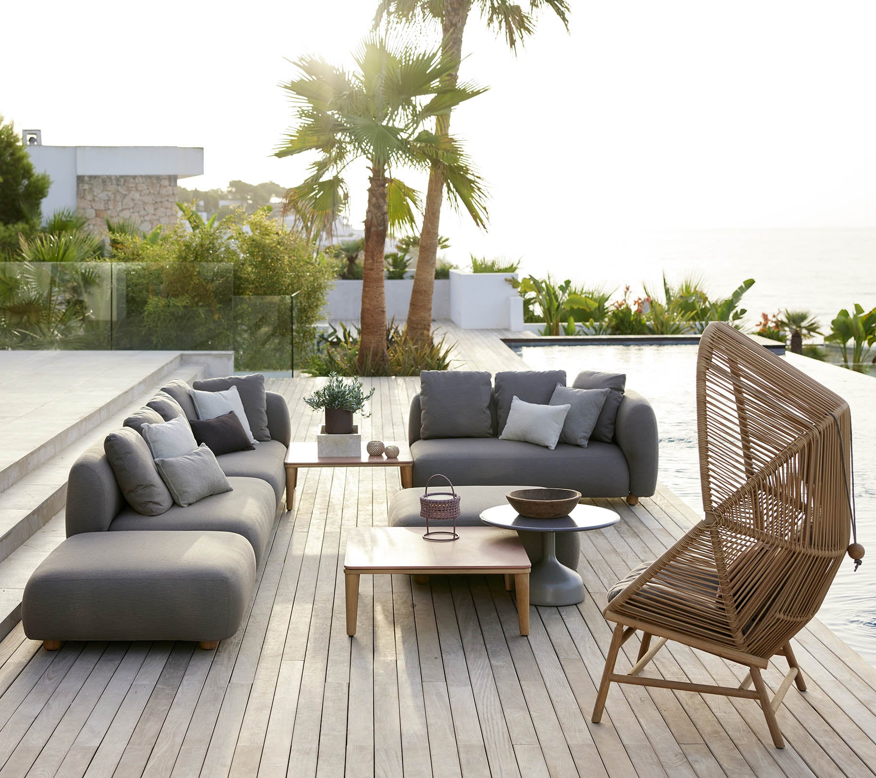 Espace de salon élégant avec des canapés gris et une chaise en rotin, complété par une table en bois et une verdure luxuriante en arrière-plan.