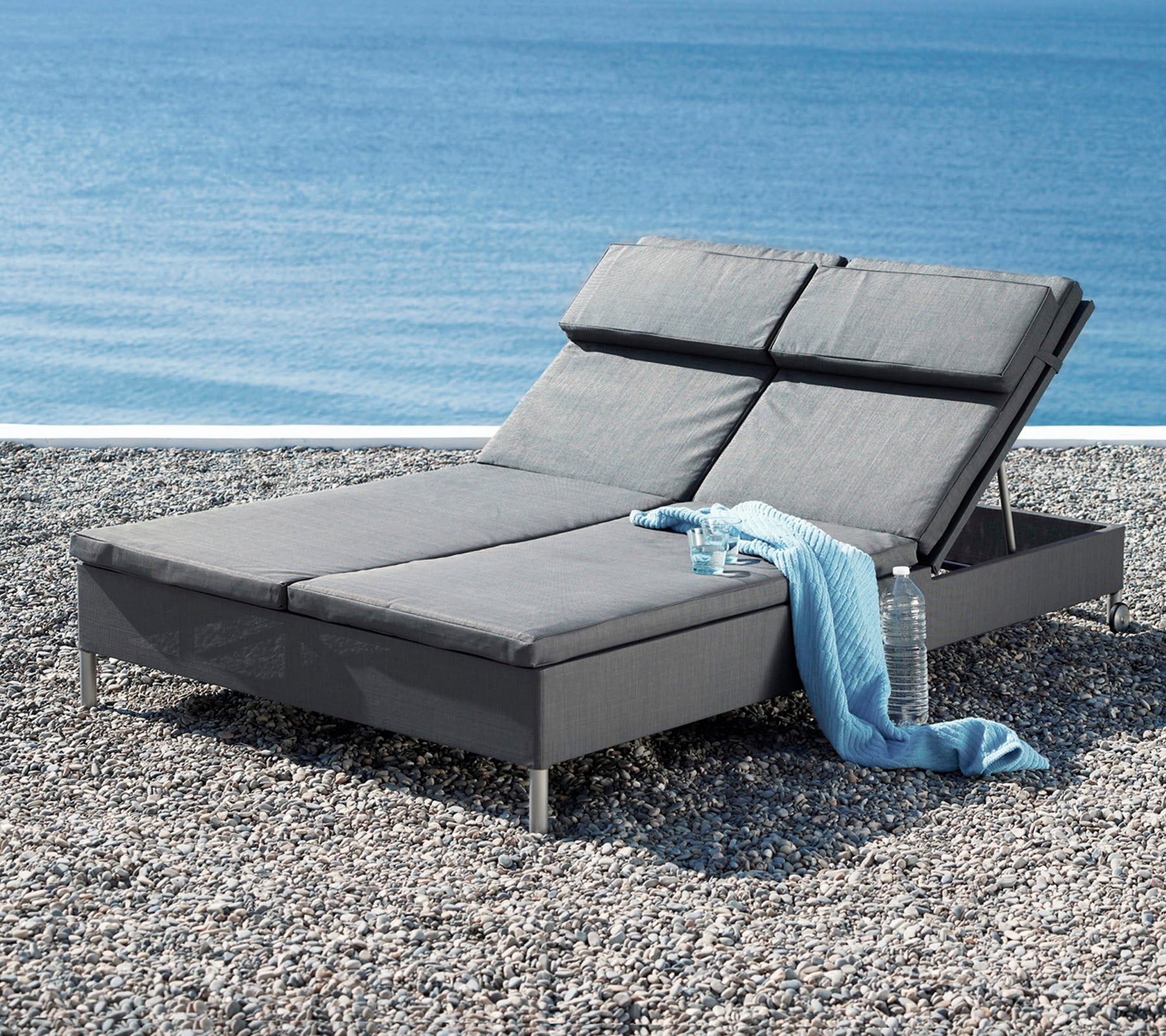Un fauteuil lounge gris élégant avec un dossier réglable, accompagné d'un doux jeté, à côté d'une vue apaisante sur l'eau.