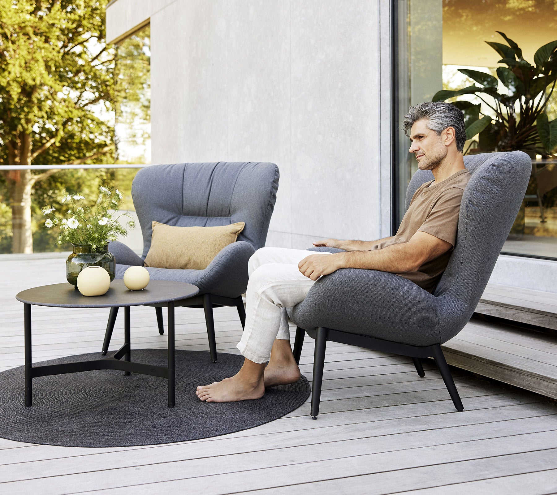 Un homme est assis dans un fauteuil confortable à côté d'une petite table avec des objets décoratifs, entouré de verdure dans un cadre serein.