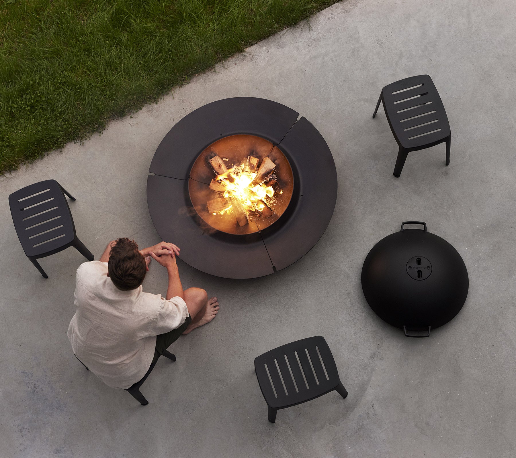 Une personne est assise près d'un foyer circulaire entouré de quatre chaises noires, profitant d'un feu chaleureux avec des bûches et un couvercle rond à proximité.